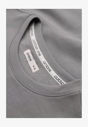 Szary bawełniany podkoszulek z okrągłym dekoltem, pokazujący wewnętrzne metki z nazwą marki "ambre" oraz rozmiarem "M".