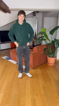 Grøn hættebluse, grå jeans og hvide sneakers. Indendørs omgivelser med gulv af træ og planter nær en orangestribet sofa.