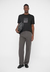 Homem em pé, vestindo uma t-shirt gráfica preta, calças cargo cinzas, tênis pretos, segurando uma grande bolsa de couro preta debaixo de um braço.