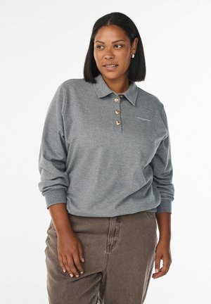 Woman wearing a grey long-sleeve polo shirt with gold buttons and brown pants, standing against a plain white background.
