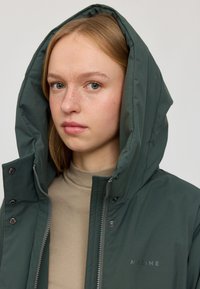 Dunkelgrüne Kapuzenjacke mit Reißverschluss und Druckknöpfen, versehen mit einem dezenten Logo auf der Brust und einem strukturierten Stoff.