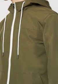 Chaqueta verde oliva con capucha, que presenta un cierre frontal blanco y cordones ajustables. Tejido suave y dos bolsillos frontales para almacenamiento.