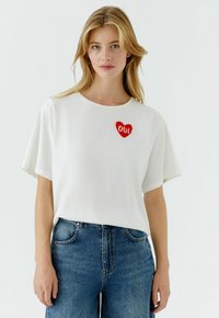 Wit katoenen T-shirt met korte mouwen, met een rood hartontwerp waarin "OUI" in het wit is geborduurd. Gecombineerd met een blauwe spijkerbroek.