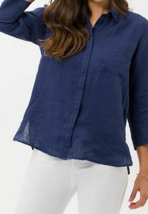 Nainen, joka pukeutunut löysään laivastonsiniseen nappipaitaan, jossa on 3/4 hihat, ja valkoisiin housuihin, poseeraa yhdellä kädellä ylhäällä.