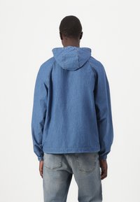 Blå denim hettegenser med elastiske mansjetter, avslappet passform og teksturert overflate. Synlige sømmer og et uformelt design.
