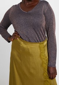 Osoba ubrana w błyszczącą, długorękawową, fioletową bluzkę włożoną do oliwkowozielonej satynowej spódnicy z koronkową lamówką, z ręką spoczywającą na biodrze.