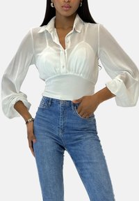 Chemisier blanc à manches longues avec un col chemise et une patte de boutons à l'avant, doté d'un corsage ajusté et de manches volumineuses. Porté avec un jean bleu.