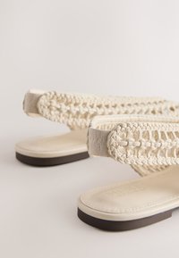 Paire de sandales couleur crème avec des brides tressées et des semelles plates noires sur un fond neutre.