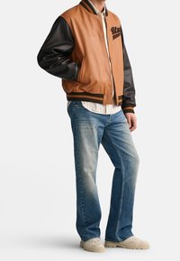 Persona vistiendo una chaqueta tipo varsity marrón y negra, una camiseta de rayas clara, jeans azules desgastados y zapatos casuales beige, de pie con las manos en los bolsillos.