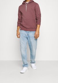 Uomo con felpa color vinaccia con tasca frontale, jeans azzurri e scarpe da ginnastica bianche in piedi davanti a uno sfondo semplice.