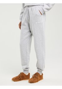 Asmuo dėvi šviesiai pilkus sportinius kelnes su elastinėmis apačiomis ir rudos spalvos zomšines sportbačius, stovintis viena ranka kišenėje.