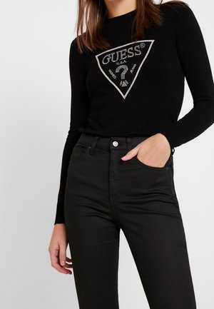 Femme portant un haut noir à manches longues avec logo Guess en strass et un jean noir taille haute, une main dans la poche, sur fond uni.