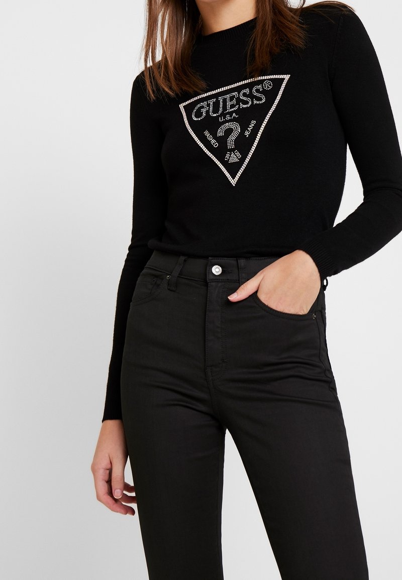 Femme portant un haut noir à manches longues avec logo Guess en strass et un jean noir taille haute, une main dans la poche, sur fond uni.