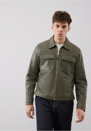Joven con chaqueta de cuero verde oliva con cremallera y bolsillos con solapa, camisa blanca y vaqueros azul oscuro sobre un fondo liso y claro.