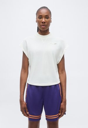 Woman wearing a white sleeveless top with a small logo and purple athletic shorts with orange stripes, standing against a plain background.