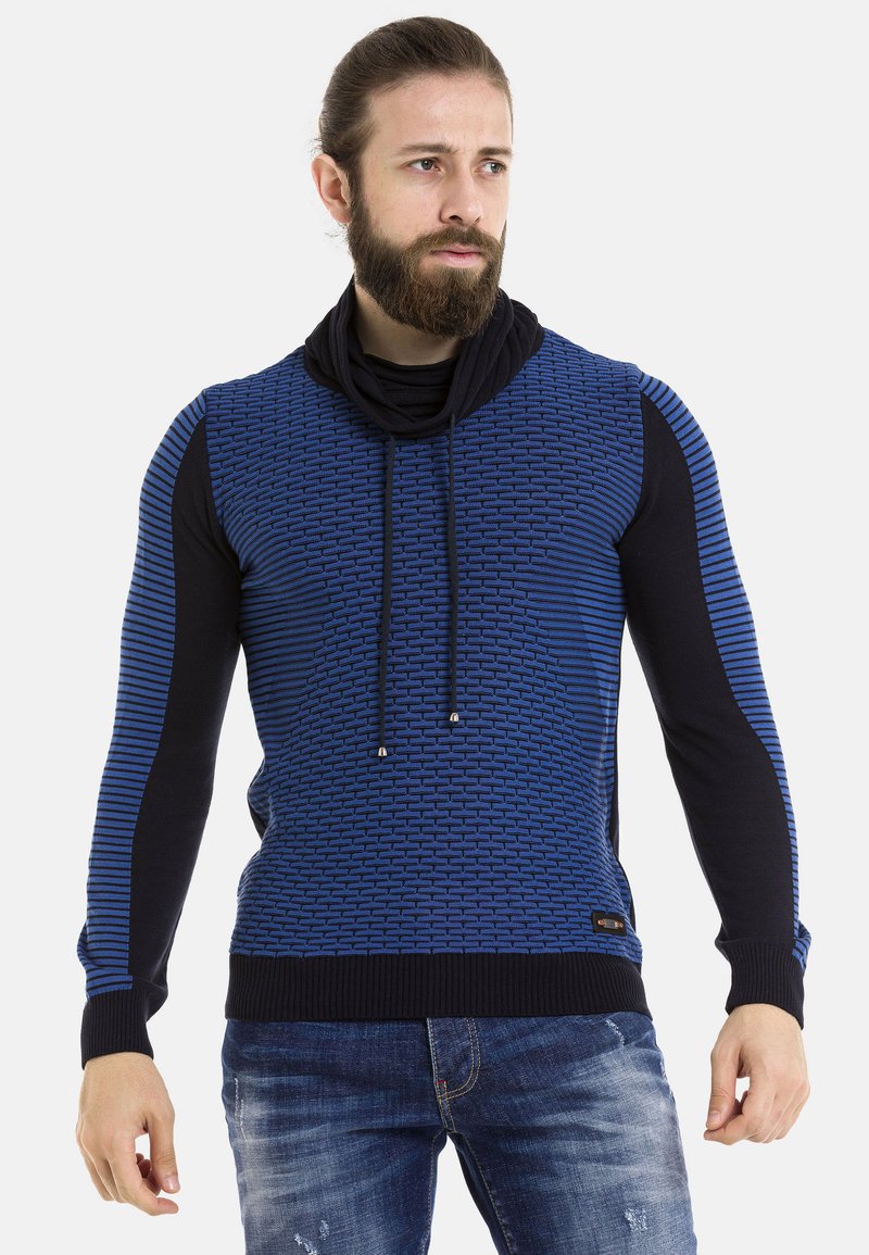 Blauer und schwarzer Pullover mit strukturiertem Muster, Kapuze und Kordelzug. Gerippte Bündchen und Saum. Getragen mit blauen, ausgebeulten Jeans.