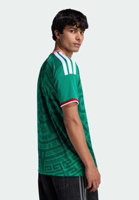 Jauns vīrietis, skatoties sāniski, valkā zaļu sporta kreklu ar baltām svītrām uz pleciem un melnas bikses.