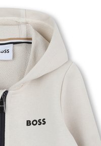 Sweat à capuche zippé couleur beige clair, avec texture côtelée et logo "BOSS" en noir. Présente une fermeture éclair bleu marine et un détail à rayures horizontales à l'intérieur du col.