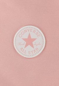 Tissu rose avec une surface texturée présentant une patch ronde blanche. Le patch comporte une étoile rose et les mots "CONVERSE ALL STAR" disposés en cercle.