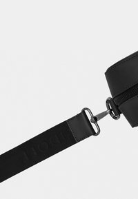 Black circular bag with a textured surface, zip closure, and adjustable strap featuring embossed lettering, attached via metallic hardware.