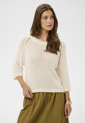 Femme aux longs cheveux bruns portant un pull en tricot crème à manches trois-quarts et une jupe vert olive, regardant sur le côté.