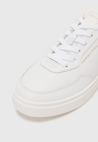 Sneaker bianco in pelle con texture liscia, punta rotonda, lacci bianchi e dettaglio del logo dorato. Suola in gomma spessa e dettagli cuciti.