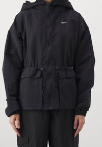 Chaqueta negra con capucha, cierre de cremallera en el frente, cintura ajustable con cordón, puños elásticos y logotipo de Nike en blanco en el pecho. Tejido ligero, diseño minimalista.