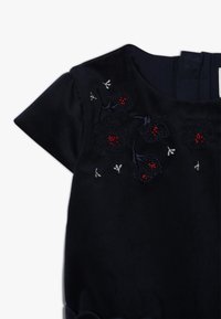 Granatowa sukienka z krótkim rękawem, z kwiatowym haftem w kolorze czerwonym i srebrnym, teksturowanym materiałem i falbaniastym wykończeniem dołu.