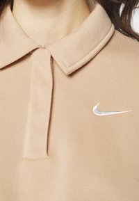 Beige polo-shirt med en vikt krage, mjuk textur och en silverfärgad Nike-logga broderad på vänster sida.