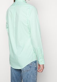 Light green striped long-sleeve shirt with a buttoned cuff and a curved hem. The fabric appears smooth, paired with denim jeans.
