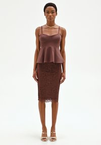Mujer con blusa marrón sin mangas con volante y falda de encaje marrón hasta la rodilla, collar dorado y sandalias de tacón abierto, de pie.