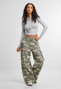 Sudadera gris de manga larga con cremallera, combinada con pantalones cargo de camuflaje de pierna ancha que cuentan con bolsillos laterales y un corte relajado.