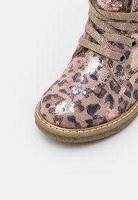 Pohled zepředu na růžovou botu s leopardím potiskem a metalickými akcenty, béžové šněrování a světle hnědá prošívaná podrážka na bílém pozadí.