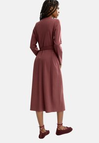 Bordowa midi sukienka z długimi rękawami i dopasowanym paskiem. Gładki materiał, dopasowana góra, rozkloszowany dół, zestawiona z bordowymi płaskimi butami na sznurówki.