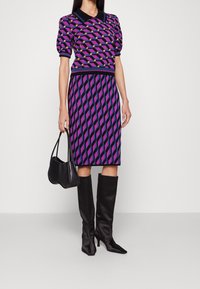 Sweterkowa sukienka z krótkimi bufiastymi rękawami, fioletowo-czarny geometryczny wzór, zestawiona z czarnymi kozakami za kolano i małą czarną torebką.