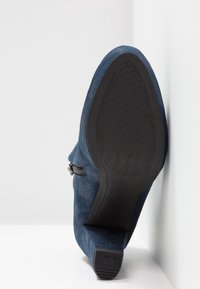 Bottine en daim bleu à la cheville, inclinée vers le haut, montrant une semelle en caoutchouc noire texturée et un petit talon bloc avec une fermeture éclair visible sur le côté extérieur.