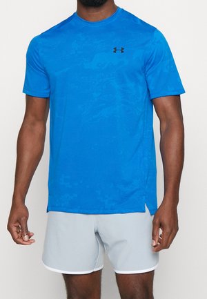 Mężczyzna w jasnoniebieskiej sportowej koszulce z krótkim rękawem z małym czarnym logo na klatce piersiowej i jasnoszarych spodenkach.