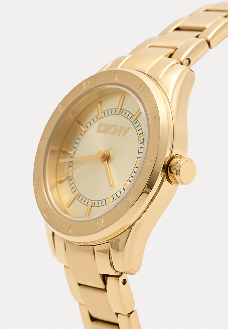 Goudkleurig DKNY-horloge met een ronde kast, gestructureerde wijzerplaat en gouden armband. Heeft minimalistische markeringen en een prominente kroon.