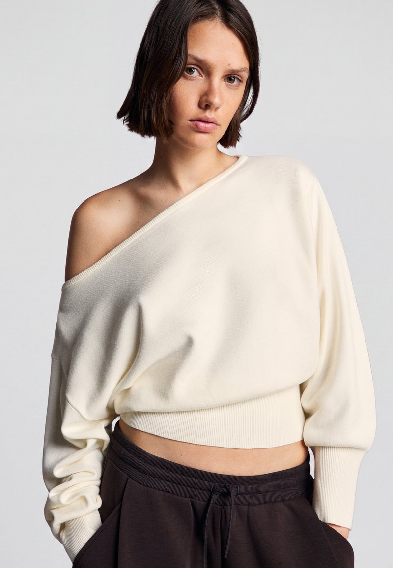 Jeune femme aux cheveux bruns courts porte un pull blanc à épaules dénudées et un pantalon foncé, posant devant un fond clair uni.