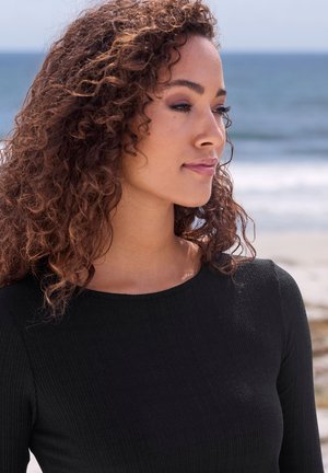 Melna ribota garām piedurknēm augšdaļa ar apaļu izgriezumu, kuru valkā persona ar cirtainiem matiem. Okeāna fons ar mīkstu fokusu viļņiem tālumā.