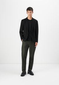 Sort blazer med notch-lapel over en sort sweater, parret med grønne bukser. Fodtøj er sorte læder loafers. Enkel, skræddersyet design.