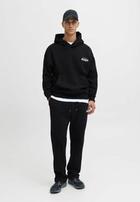 Sort hættetrøje med frontlomme og logo, parret med sorte sweatpants. Tilbehør inkluderer en matchende sort kasket og mørke sneakers.