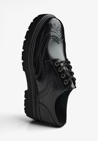 Scarpa brogue in pelle nera con suola spessa e strutturata, perforazioni decorative e un tradizionale sistema di allacciatura. Presenta una finitura liscia e lucida.