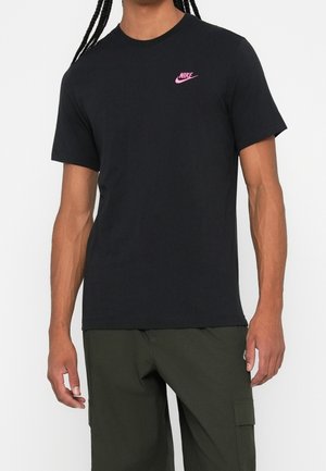 Osoba nosząca czarną koszulkę Nike z różowym logo na piersi i ciemnozielone spodnie typu cargo, stojąca z ramionami swobodnie opuszczonymi wzdłuż ciała.