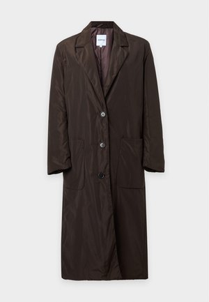 Femme portant un long manteau marron foncé boutonné par-dessus un haut bordeaux avec un pantalon large beige et des chaussures foncées, debout devant un fond uni.