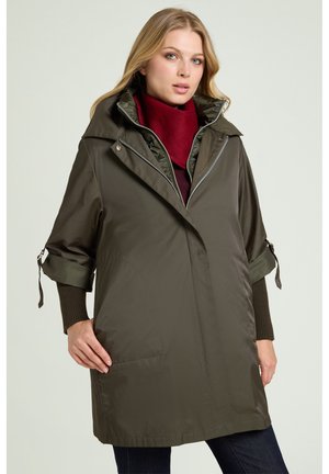 Woman wearing a dark green oversized coat with rolled-up sleeves and a maroon scarf, standing against a plain light background.