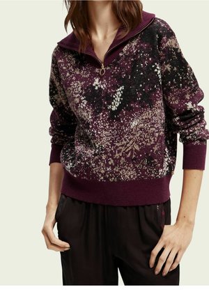 Femme portant un pull à demi-fermeture éclair violet à motifs floraux et un pantalon noir, les mains détendues le long du corps, sur un fond clair.