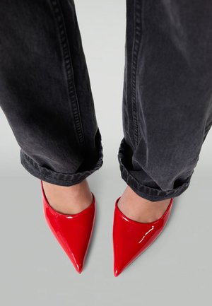 Red patent leather high heels with pointed toes, paired with rolled-up dark gray jeans, showcasing a sleek design and glossy finish.