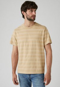 Beżowa koszulka z krótkim rękawem w poziome białe i jasnopomarańczowe paski. Okrągły dekolt, materiał bawełniany, luźny krój. Noszona z niebieskimi dżinsami.