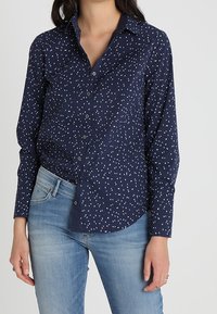 Chemise bleu marine à boutons avec un col à revers et un motif de petits points blancs éparpillés. Manches longues avec poignets repliés. Portée avec un jean bleu clair.